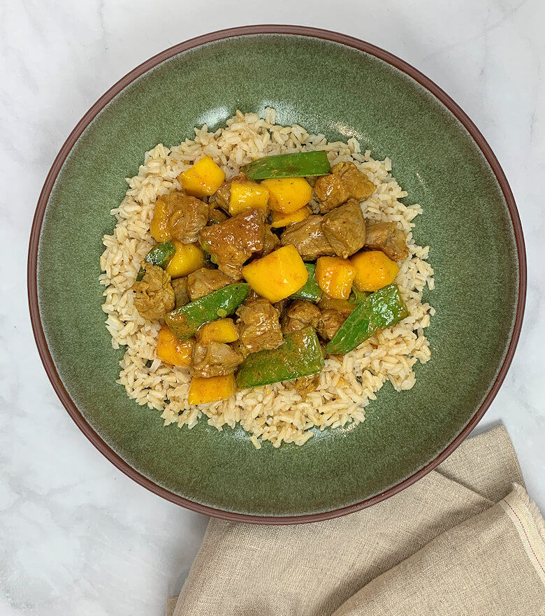 Green bowl with Curried Pork in Coconut Sauce with Mango & Snow Peas on a bed of brown rice.