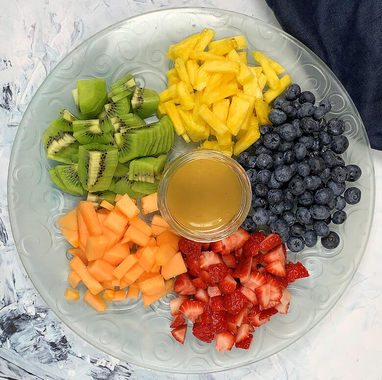 Glass plate with 5 different fruits cut up in bite size pieces and jar of dressing in the middle